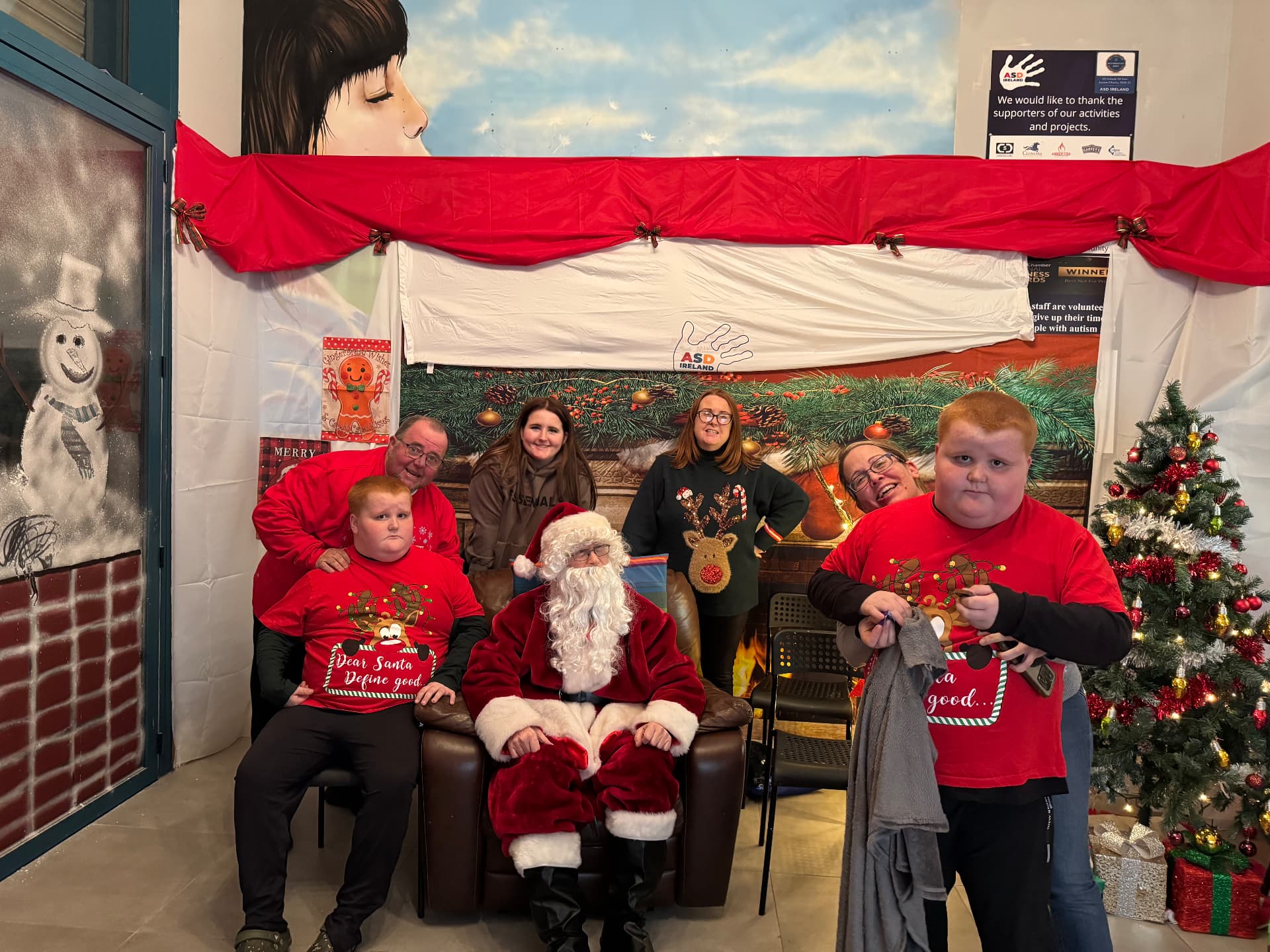 Families and volunteers with Santa at the ASD Ireland Christmas party, beside a decorated tree and festive backdrop.