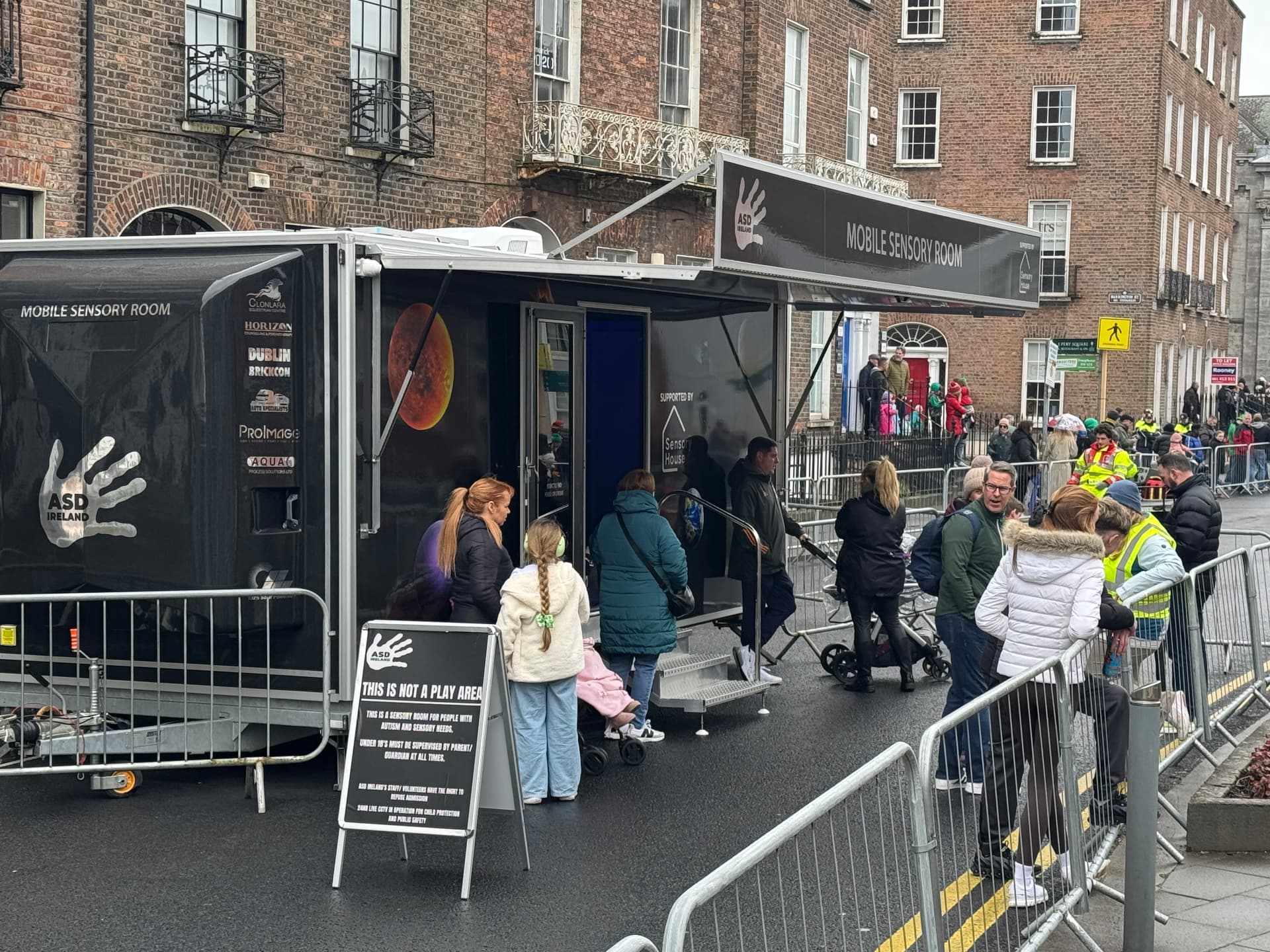 Families queueing to visit the ASD Ireland mobile sensory room on a city street, with a sign explaining it is a sensory room, not a play area.