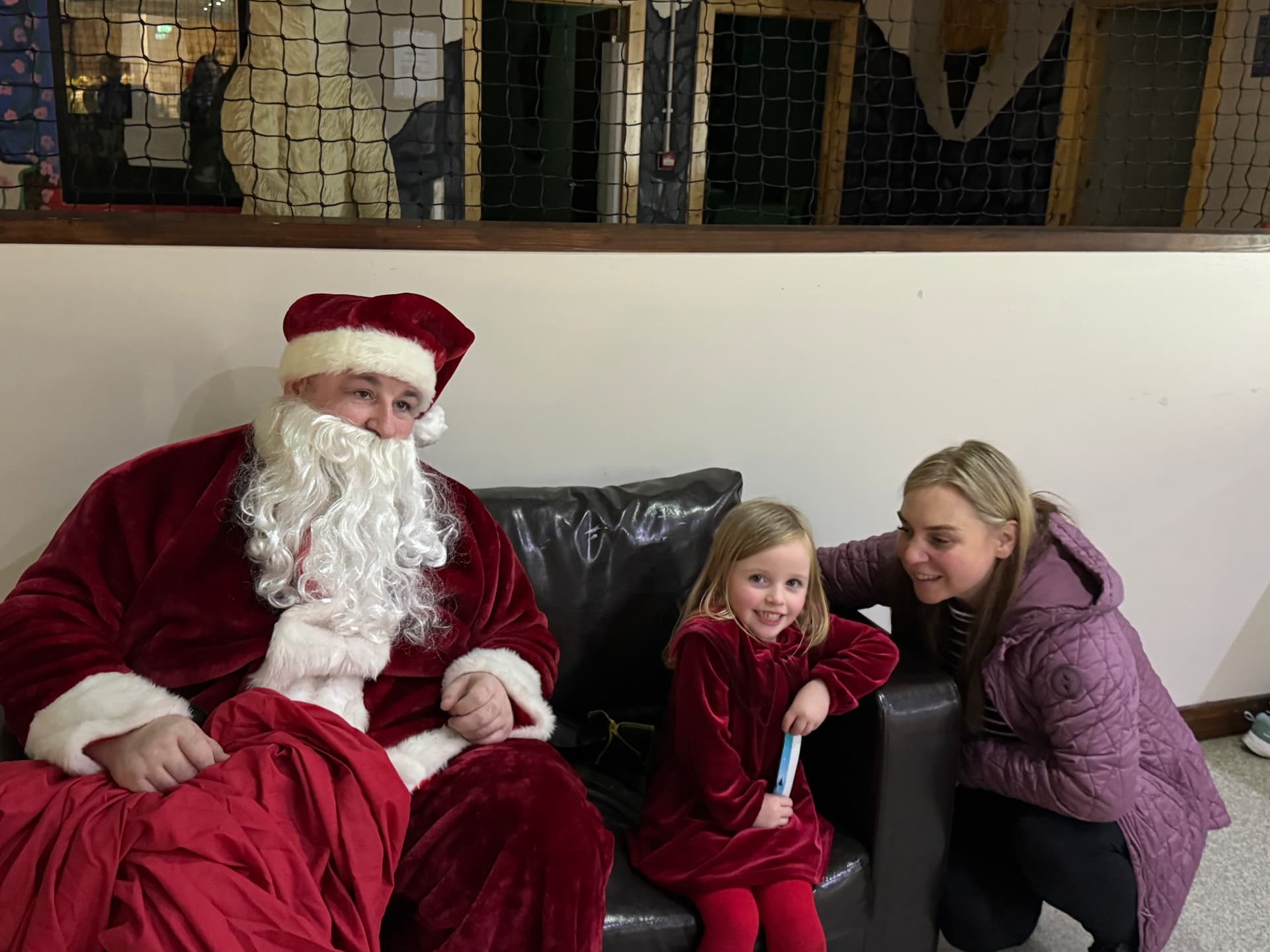 Santa sitting with a smiling child in a red velvet dress and her mum at the ASD Ireland Christmas party.