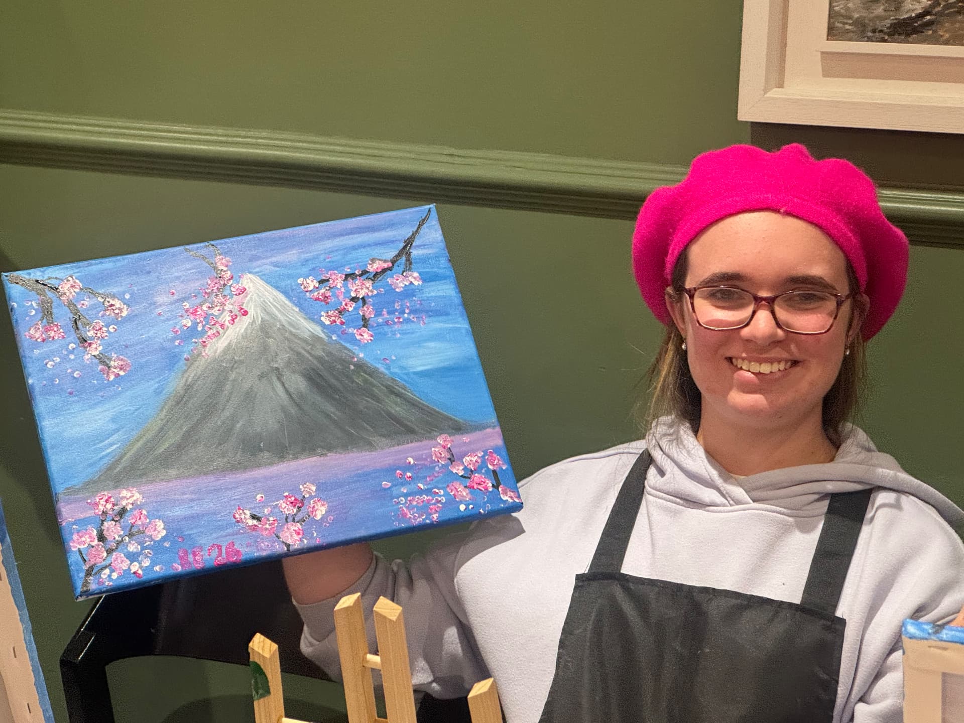A young woman in a pink beret holding up her finished painting of Mount Fuji with cherry blossoms at the Newry social group.