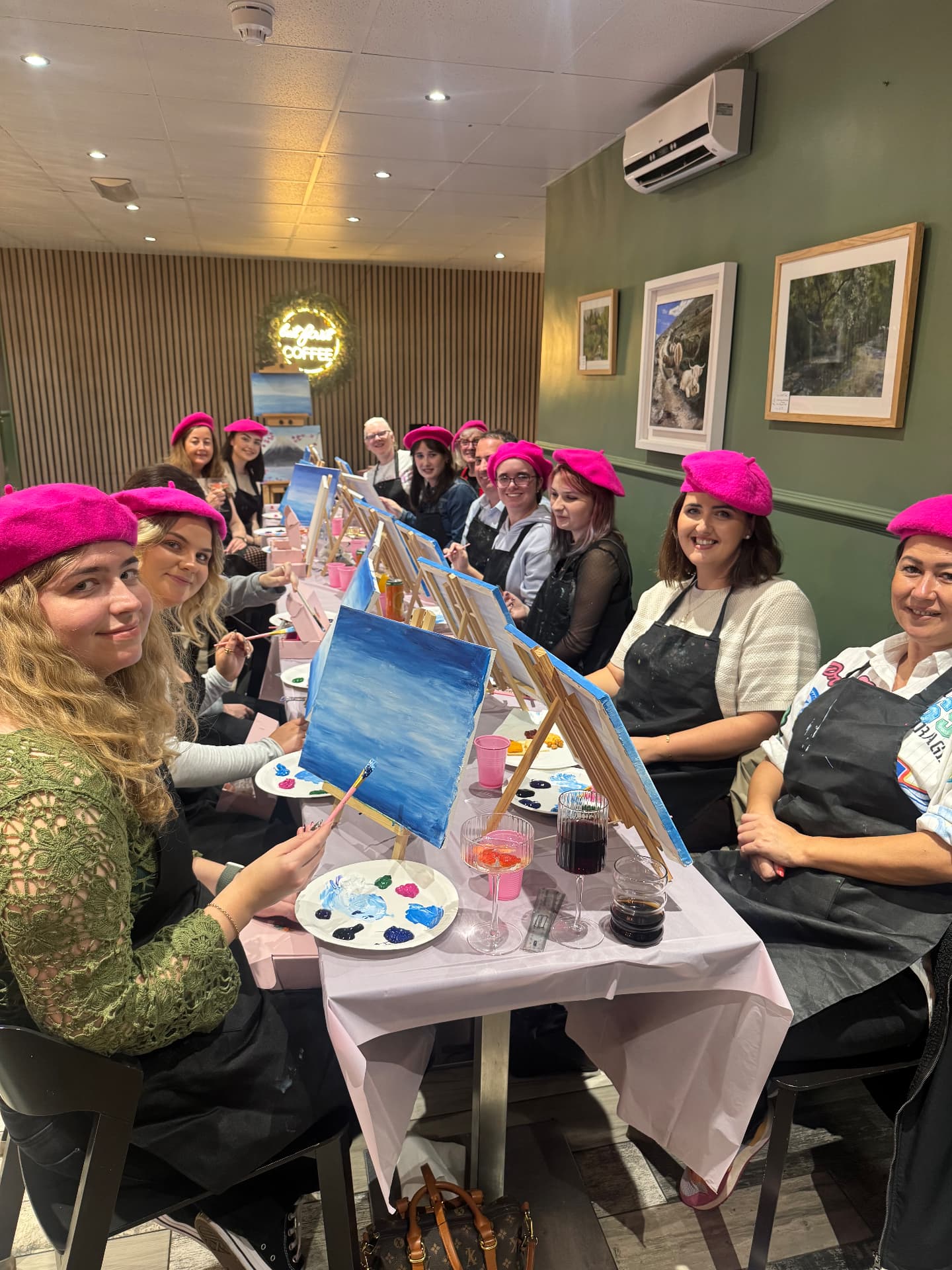 The whole Newry social group sitting at a long table with easels, pink berets and painting supplies, smiling together.