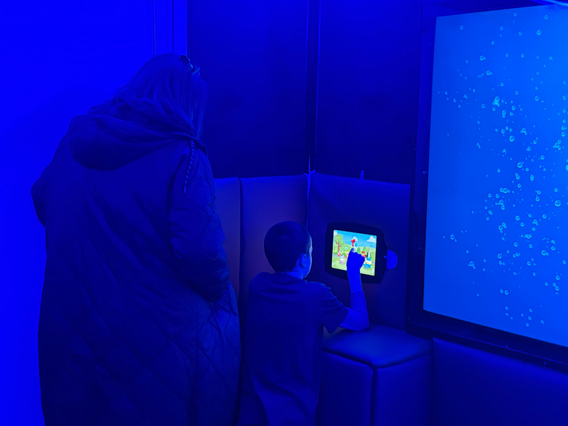 Inside the mobile sensory room: a parent and child beside a glowing blue bubble wall, the child using an interactive touch panel.