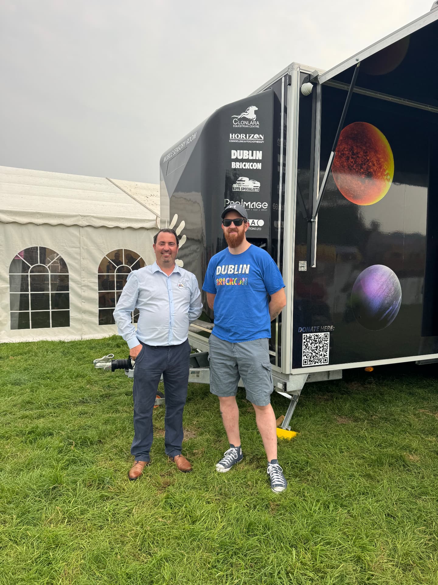 Two men standing beside the ASD Ireland mobile sensory room at an event, with supporter logos and a donation QR code on the trailer.